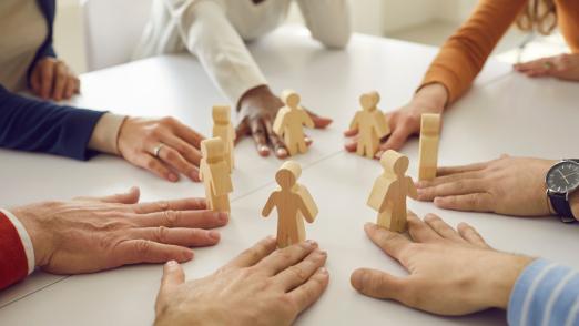 Team of multiracial business people and workmates sitting around white office table put little wooden human figures in circle as symbol of group, community, help, collaboration and teamwork